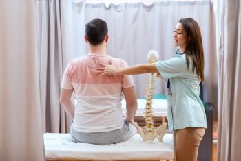 Beautiful female doctor with brown hair holding spine model and touching patient on his backs.